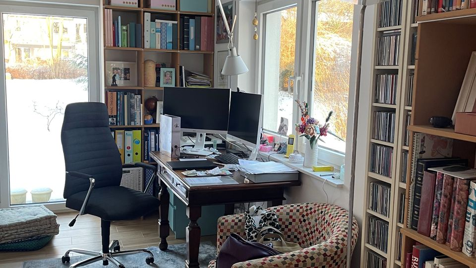 Helles Arbeitszimmer mit Holzfußboden, Schreibtisch mit zwei Monitoren und Schreibtischlampe sowie einem Drehstuhl auf einem Teppich. An den Wänden stehen hohe Regale mit Büchern und CDs; große Fenster und eine Glastür öffnen den Blick in einen Garten.