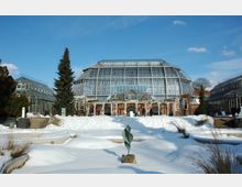 Großes Gewächshaus aus Glas und Metall in einem botanischen Garten, umgeben von verschneiten Landschaften. Im Vordergrund stehen eine Skulptur und schneebedeckte Pflanzenbeete, während sich rechts und links weitere gläserne Gewächshausstrukturen erstrecken.