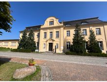 Ein historisches, zweistöckiges Gebäude mit gelber Fassade, symmetrischer Gestaltung und einem geschwungenen Giebel über dem Eingangsbereich. Vor dem Gebäude stehen mehrere Tannenbäume, ein gepflasterter Weg führt daran vorbei, und im Vordergrund befindet sich ein Stein mit einem Blumentopf.