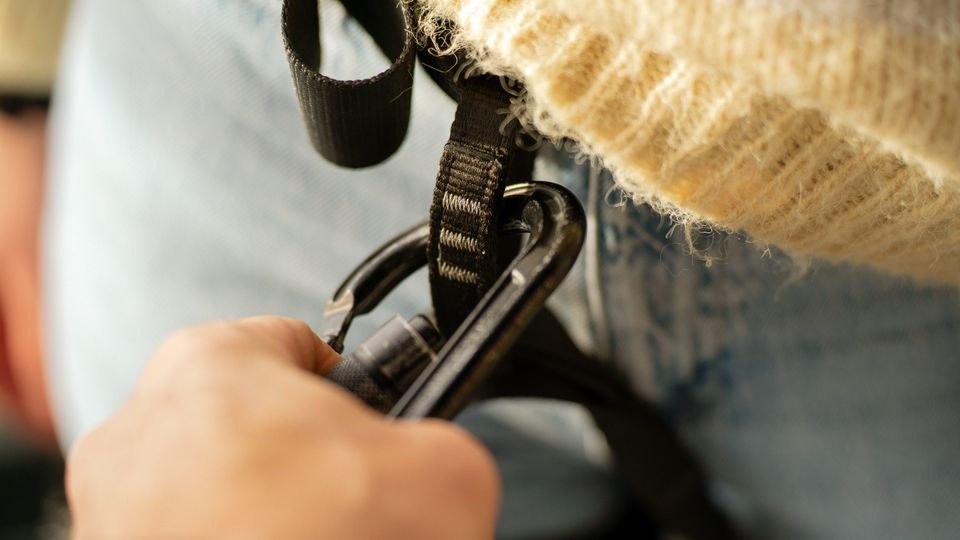 Nahaufnahme von schwarzen Karabinern und Gurtbändern eines Klettergurts, die an einer Person mit Jeans und einem beigen Strickpullover befestigt sind. Der Hintergrund ist unscharf und zeigt keine klar erkennbaren Umgebungselemente.