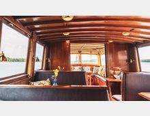 Innenraum eines Bootes mit Holzverkleidung, gepolsterten Sitzbänken und Tischen, dekoriert mit kleinen Blumenvasen. Die Fenster bieten einen Blick auf das Wasser und eine Landschaft am Horizont.