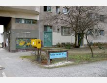 Wohnhausfassade eines grauen Gebäudes mit mehreren Fenstern und einer begrünten Fläche davor, auf der ein gelber Briefkasten der Deutschen Post steht. Neben einem Baum befindet sich ein Schild mit der Aufschrift „Waschhaus Aroser Allee 153 a, b, c“.