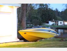 Gelbes Motorboot, das auf einem Rasen nahe einem kleinen Fluss oder Kanal steht. Im Hintergrund sind Bäume und ein weißes Wohnhaus mit Garten sichtbar.