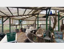 Überdachte Terrasse mit Holzboden, unterschiedlichem Gartenmobiliar und Möbeln, teils gestapelt. Die Konstruktion ist aus Holz und Metall, das Dach besteht aus durchscheinenden Wellplatten. Im Hintergrund sind Fenster, ein Zaun und Pflanzen sichtbar.