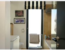 Kleines Badezimmer mit moderner Einrichtung, bestehend aus einer Toilette, einer Badewanne und einem Handtuchhalter vor einer bodentiefen, mattierten Fensterscheibe mit einer schwarz-weiß gestreiften Jalousie. An den Wänden hängen zwei gerahmte Bilder, und auf dem rechten Badewannenrand stehen Hygieneartikel.