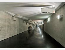 Langer, moderner Flur mit grauen Betonwänden, schwarzem glänzendem Boden und einer abgehängten Decke mit Beleuchtung. An der rechten Wand befinden sich Türen zu den Umkleidekabinen, und ein Schild zeigt den Bereich „Umkleiden 14–17“ an.