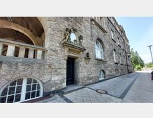 Sandsteinfassade eines historischen Gebäudes mit einem Eingang zum "Ratskeller", verziert mit einer Figurengruppe und einem ovalen Fenster darüber. Rechts entlang der Fassade befinden sich hohe, bogenförmige Fenster; der gepflasterte Gehweg führt an der Straße entlang.