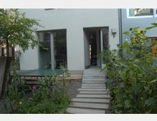 Moderner Hinterhof mit einer Terrasse, die mit einer Tischtennisplatte ausgestattet ist, umgeben von bepflanzten Beeten mit Blumen und Sonnenblumen. Eine Tür und bodentiefe Fenster führen in ein Wohnhaus mit heller, schlichter Fassade.