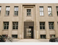 Fassade eines mehrstöckigen Gebäudes aus beigefarbenem Stein mit rechteckigen Fenstern und einem zentralen Eingang. Über dem Eingang steht "Quartier 110", und davor befinden sich Fahrräder und geparkte Autos.