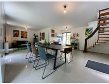 Modernes Wohnzimmer mit offenem Essbereich, einem großen Esstisch und bunten Stühlen. Im Hintergrund sind eine graue Couch, abstrakte Wandbilder, eine freistehende Holztreppe und eine Glastür mit Blick nach draußen sichtbar.