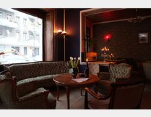 Gemütlicher Innenraum eines Cafés oder Salons mit antiken Möbeln, darunter grüne Sessel, ein runder Holztisch und ein Sofa. Im Hintergrund befinden sich eine alte Radioanlage, ein Kerzenleuchter und eine floral gemusterte Wandtapete, während große Fenster den Blick auf eine Stadtstraße ermöglichen.