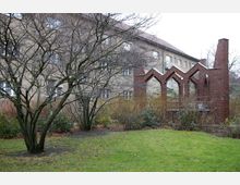 Grüne Gartenfläche mit kahlen Bäumen, im Hintergrund ein Wohngebäude mit Ziegeldach und brauner Fassade. Rechts steht eine Ziegelkonstruktion mit bogenförmigen Durchgängen und einem aufragenden Schornstein.