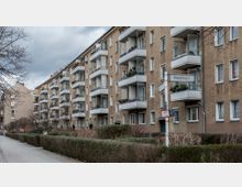 Mehrstöckiges Wohngebäude aus beige-braunem Putz mit zahlreichen Balkonen, die mit Pflanzen dekoriert sind. Im Vordergrund ein Fußweg, gesäumt von Hecken und kahlen Bäumen, sowie ein Straßenschild mit der Aufschrift "Binzstraße".