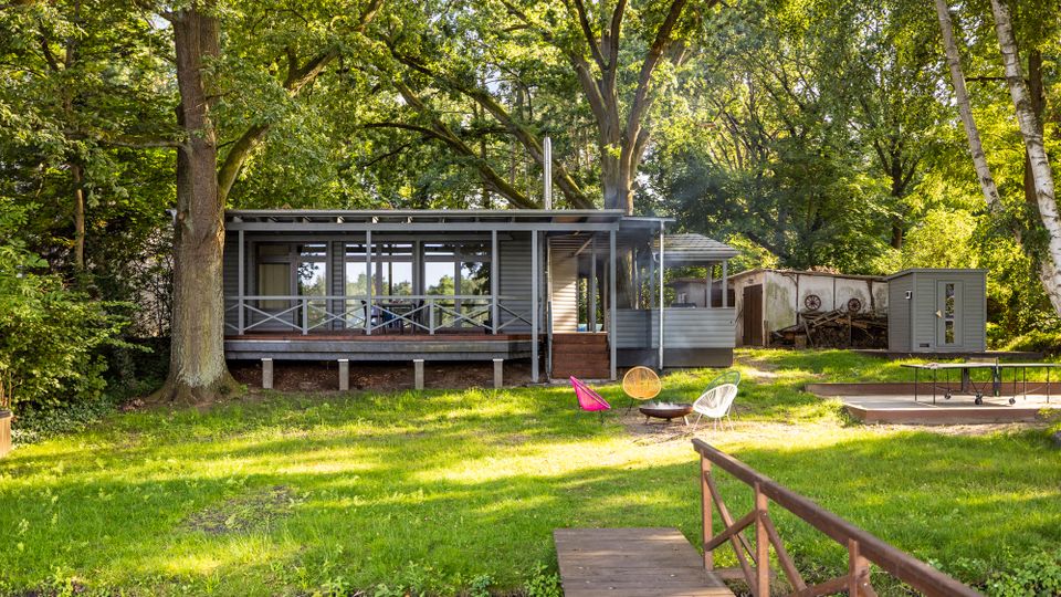 Modernes, einstöckiges Holzhaus mit großer überdachter Veranda und Glasfront in einem lichtdurchfluteten Waldgrundstück mit hohen Laubbäumen. Davor liegt eine Rasenfläche mit Feuerstelle und Sitzschalen; rechts sind ein Holzsteg über einen Graben, ein kleiner Schuppen sowie eine gepflasterte Fläche mit Tischtennisplatte zu sehen.