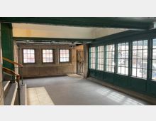 Innenraum einer Fußgängerpassage, vermutlich in einer historischen Bahnstation, mit grünen Metallrahmen und großen Fenstern, durch die die umliegenden Gebäude und Straßen sichtbar sind. An der linken Seite führt ein Treppengeländer aus Metall mit einem Holzhandlauf nach oben.