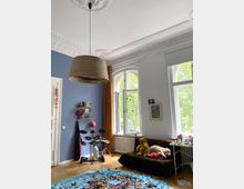 Ein Kinderzimmer mit hohen Decken und dekorativen Stuckverzierungen. Der Raum hat eine blaue Wand, große Bogenfenster mit Blick ins Grüne, ein Sofa mit Kuscheltieren, ein elektronisches Schlagzeug und einen Boden, der teilweise mit bunten Legosteinen bedeckt ist.