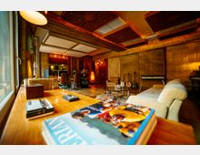 Aufnahme eines gemütlichen Musikstudios mit Holzwänden und -möbeln. Im Raum befinden sich Instrumente wie Gitarren und ein Keyboard, zwei Schreibtische mit Monitoren, sowie eine Sitzgruppe mit Sofa, Couchtisch und dekorativer Beleuchtung.