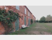 Backsteingebäude mit grünen Holztüren und Fenstern, umgeben von Rosensträuchern und einer Rasenfläche. Im Hintergrund stehen Bäume, Bänke und ein kleiner Pavillon auf dem weitläufigen Grundstück.