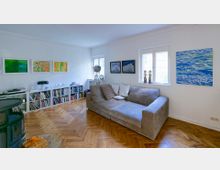 Wohnzimmer mit hellen Wänden, Parkettboden und großen Fenstern. Eine graue Eckcouch steht vor einer Wand mit farbenfrohen Gemälden, daneben ein Regal mit Büchern und Dekorationsgegenständen sowie ein runder Kamin im Vordergrund.