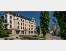 Eckgebäude mit heller Fassade und Dachgauben, teilweise mit Balkonen, sowie einem markanten runden Turm mit Kuppel auf der rechten Seite. Vor dem Gebäude befinden sich ein gepflasterter Platz, einzelne Bäume und Grünflächen.