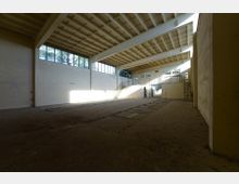 Ein leerer, rechteckiger Innenraum mit hohen Decken, sichtbaren Trägerkonstruktionen und großen Fenstern mit rechteckigen Glasflächen an der oberen Wandzone. Der Raum hat einen staubigen Betonboden, beleuchtete Wandflächen durch Sonnenlicht und eine Treppe, die zu einer oberen Ebene führt.