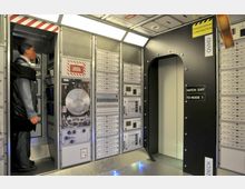 Innenraum einer Raumstation mit technischen Kontrolltafeln und Schaltflächen an den Wänden. Ein schmaler Durchgang mit der Aufschrift „Hatch Exit to Node 1“ führt in einen angrenzenden Bereich, und eine lebensgroße Puppe steht links in einer Nische.