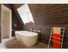 Modernes Badezimmer mit freistehender Badewanne vor einer schrägen Wand, die mit dunklen Fliesen verkleidet ist. Auf dem Boden befinden sich Kieselsteinfliesen, und ein Fenster lässt Tageslicht herein; daneben steht ein Handtuchhalter mit einem roten und einem beigen Handtuch.