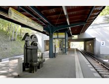 U-Bahn-Station mit Überdachung, Sitzbank in moderner Skulptur-Optik, gelbem Fahrkartenautomaten und Hinweisschild „Dahlem-Dorf“; daneben Gleis 2 mit erhöhtem Bahnsteig und angrenzender grüner Böschung.