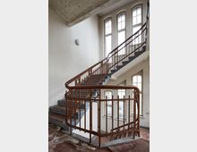 Treppe in einem Altbau mit geschwungenem, braunem Metallgeländer und grauen Stufen. Große Fenster mit Sprossen lassen viel Tageslicht in den Treppenraum.