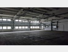 Großer, leerer Industrieinnenraum mit Betonboden, weißen Wänden und einer Decke mit sichtbaren Trägern. Mehrere Fensterreihen lassen diffuses Tageslicht herein, während technische Installationen und Rohrleitungen an den Wänden und der Decke sichtbar sind.