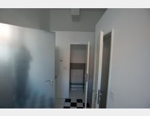 Ein Innenraum mit weiß gestrichenen Wänden, einer halb geöffneten Milchglastür auf der linken Seite und mehreren weißen Innentüren. Der Boden besteht aus schwarz-weiß karierten Fliesen, und im Hintergrund ist eine Wand mit einem schwarz-weiß gemusterten Fliesenspiegel sichtbar.