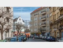 Straßenszene in einer urbanen Wohngegend mit mehrstöckigen Altbau-Wohnhäusern in hellen Farben, teilweise mit Balkonen. Im Hintergrund sind das Gebäude mit der Aufschrift "Schöneberger Tor" und ein Metallgerüst sichtbar. Autos parken auf beiden Seiten der Kopfsteinpflasterstraße, und unten befinden sich kleine Geschäfte und Restaurants.