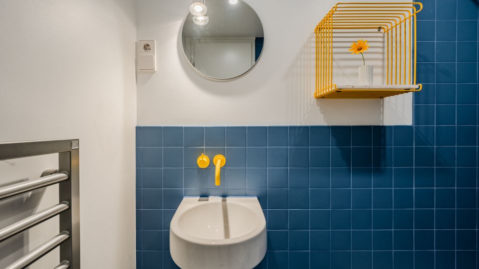 Modernes Badezimmer mit wandhängendem weißem Waschbecken vor blauen Fliesen und gelben Armaturen. Darüber hängen ein runder Spiegel mit Lampe sowie ein gelbes Wandregal; links ist ein Handtuchheizkörper zu sehen.