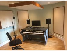 Ein Musikstudio mit Schreibtisch, auf dem ein digitales Keyboard, ein Computerbildschirm und Lautsprecher stehen. Eine Akustikverkleidung ist an den Wänden montiert, und ein Mikrofonständer steht neben einem Bürostuhl auf einem Holzboden.