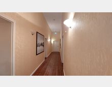 Langer Flur mit beige gestrichenen Wänden und Holzfußboden, beleuchtet durch Wandlampen. An den Wänden hängen gerahmte Bilder, und am Ende des Flurs befindet sich eine weiße Tür.