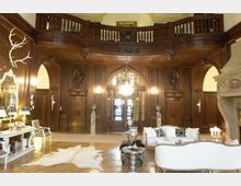 Eleganter Salon mit Holzvertäfelung an den Wänden, hohen Doppeltüren in der Mitte und einer umlaufenden Empore. Im Raum befinden sich weiße Sofas, ein Kamin auf der rechten Seite und ein großflächiger Holzboden mit einem hellen Fellteppich.