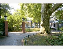 Ein kleiner Park mit gepflastertem Weg, umgeben von Zäunen aus grünem Gitterwerk. Backsteinpfeiler mit alten Laternen rahmen einen Eingang, während im Vordergrund ein großer Baum und Beete mit Pflanzen zu sehen sind.