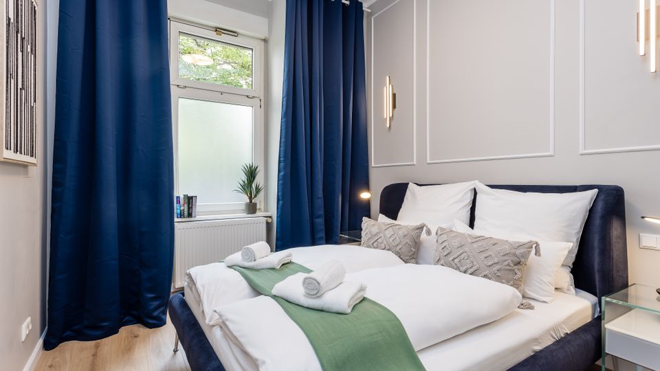 Modernes Schlafzimmer mit Doppelbett in weißer Bettwäsche, blauem Polsterbettgestell und grünem Bettläufer; an den Wänden sind Zierleisten und schlichte Wandlampen. Ein Fenster mit dunkelblauen Vorhängen, Heizkörper darunter sowie ein kleiner Beistelltisch aus Glas ergänzen den Raum.