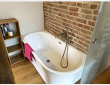 Freistehende weiße Badewanne mit moderner Armatur vor einer Ziegelsteinwand. Daneben steht ein offenes Regal aus Holz mit Badutensilien, und der Boden besteht aus Holzplanken.