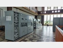 Ein historischer Kontrollraum mit grauen Schalttafeln, analogen Anzeigen und Steuerknöpfen. Der Raum hat eine hohe Decke, große Fenster, die viel Tageslicht hereinlassen, und einen gemusterten Fliesenboden.