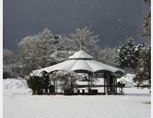 Ein schneebedeckter Pavillon steht inmitten einer verschneiten Landschaft mit kahlen, schneebedeckten Bäumen im Hintergrund. Der Himmel ist dunkelgrau, und fallender Schnee ist sichtbar.