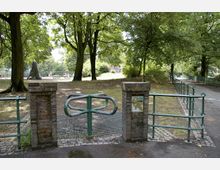 Eingang zu einem Spielplatz in einem Park, umgeben von Bäumen. Der Zugang ist durch eine grüne Metall-Drehschranke und Zäune mit gemauerten Pfeilern begrenzt; ein Schild weist auf den Kinderspielplatz hin.