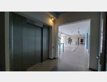 Innenansicht eines Gebäudes mit Blick auf einen Flur und einen Aufzugsbereich. Der Flur ist hell beleuchtet, mit gefliesten Böden, blauen Wänden und großen Fenstern; Pflanzen und Sitzmöbel sind als Dekoration vorhanden.