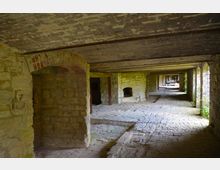 Ein langer, steinerner Gewölbegang mit grobem Mauerwerk, niedriger Decke und Boden aus ungleichmäßigen Steinplatten. Im Hintergrund sind Öffnungen, kleine Bogenfenster und ein Kamin sichtbar, durch die Tageslicht einfällt.