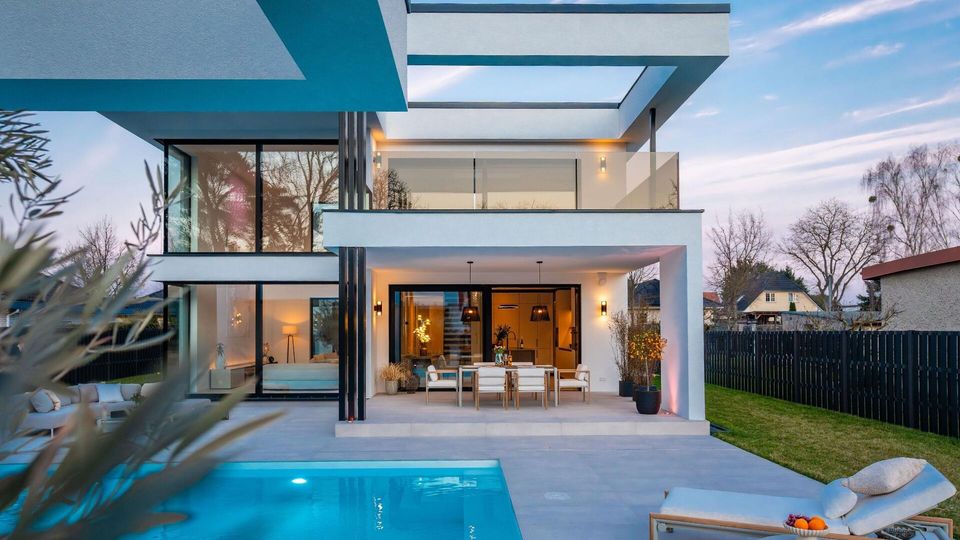 Modernes, zweigeschossiges Wohnhaus mit klaren, kubischen Formen, großen Glasfronten und überdachter Terrasse mit Esstisch. Davor liegen eine gepflasterte Poolterrasse mit rechteckigem Swimmingpool und Liege, umgeben von Rasenfläche und dunklem Zaun.