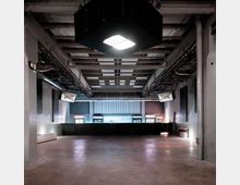 Ein leerer, moderner Veranstaltungssaal mit Betonfußboden und sichtbarer Deckenstruktur. An der Decke hängt ein großer quadratischer Lichtkasten, und links und rechts sind Lautsprecher befestigt; im Hintergrund befindet sich eine erhöhte Bühne mit niedriger Rückwand und Fenstern.