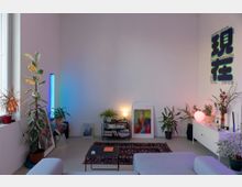 Modernes Wohnzimmer mit großer weißer Wand, vielen Zimmerpflanzen und indirekter Beleuchtung durch eine blaue LED-Röhre und eine runde Tischlampe. Vor der Wand stehen ein niedriges Regal mit Büchern, gerahmte Bilder am Boden, ein gemusterter Teppich mit Couchtisch sowie ein Sideboard mit Deko und Poster.