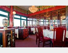 Asiatisch eingerichtetes Restaurant mit roten Holzmöbeln und weißen Tischdecken. Die Wände sind reich verziert mit roten und floralen Ornamenten, und eine große Kristallleuchte hängt von der Decke; Vasen und dekorative Schränke verstärken das traditionelle Dekor.