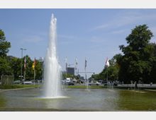Ein großer Springbrunnen mit hohem Wasserstrahl in der Mitte eines flachen Beckens, umgeben von Bäumen, wehenden National- und Länderflaggen sowie einer Straße mit vorbeifahrenden Autos im Hintergrund.