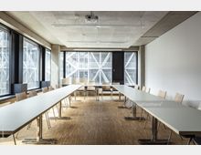 Moderner Konferenzraum mit großen Fenstern, die Blick auf eine stählerne Fassadenkonstruktion erlauben. Der Raum hat helle Tische in U-Form, einfache Holzstühle und einen Boden aus hellem Parkett.